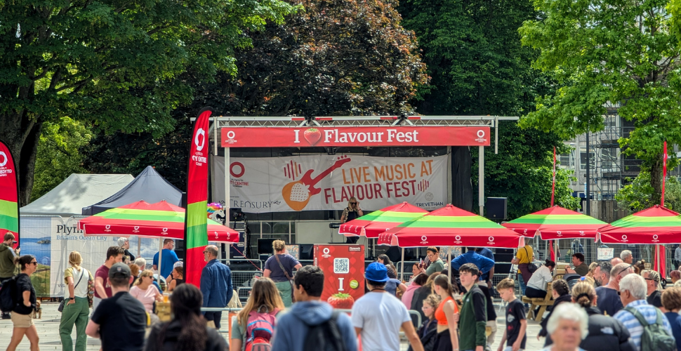 Crowds enjoy an outdoor “I Love Flavour Fest” live music stage in a sunny city square, with red and green parasols, food stalls and leafy trees creating a busy, festival atmosphere.